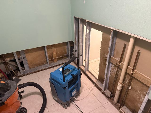 Bathroom with a blue dehumidifier and a wet-vac on the floor and the wall cavity opened.